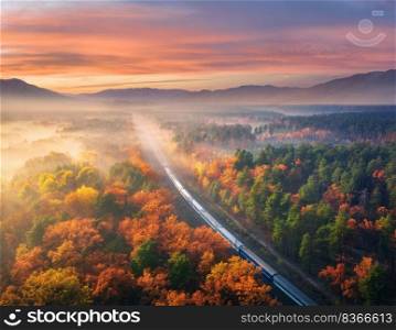 Aerial view of passenger train in beautiful forest in fog at sunset. Autumn landscape with railroad, foggy trees, trail and colorful sky with clouds. Top view of moving train in fall. Railway station. Aerial view of passenger train in autumn forest in fog at sunset