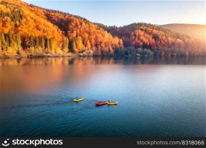 Aerial view of people on floating colorful boats on blue lake in mountains with red forest at sunset in autumn. River in carpathian mountains in fall in Ukraine. Landscape. Top view of canoe. Tourism