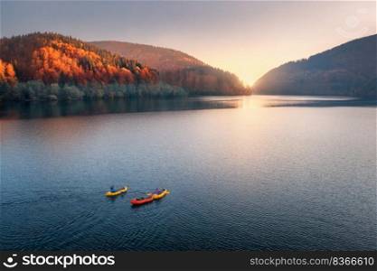 Aerial view of people on floating colorful boats on blue lake in mountains with red forest at sunset in autumn. River in carpathian mountains in fall in Ukraine. Landscape. Top view of canoe. Travel. Aerial view of people on boats on blue lake in mountains