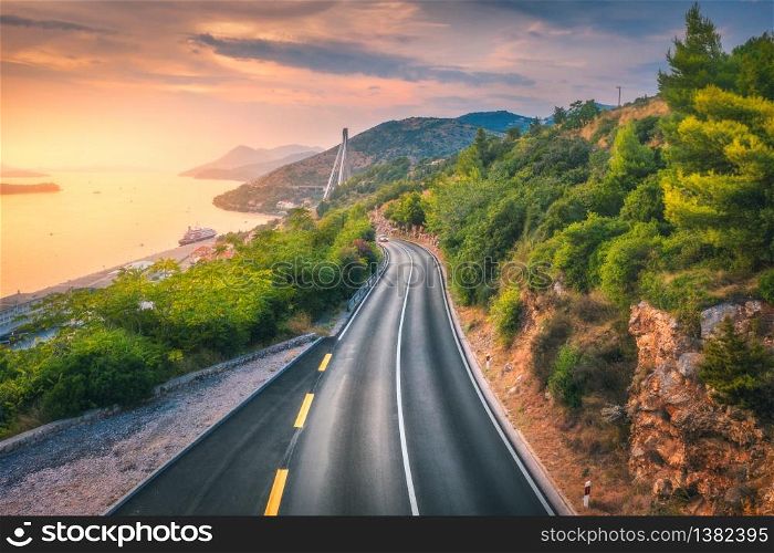 Aerial view of perfect mountain road and beautiful green forest at colorful sunset in summer. Dubrovnik, Croatia. Top view of road, sea, mountain, sky. Landscape with highway, sea coast, gold sunlight. Aerial view of perfect mountain road and beautiful green forest