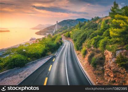 Aerial view of perfect mountain road and beautiful green forest at colorful sunset in summer. Dubrovnik, Croatia. Top view of road, sea, mountain, sky. Landscape with highway, sea coast, gold sunlight. Aerial view of perfect mountain road and beautiful green forest