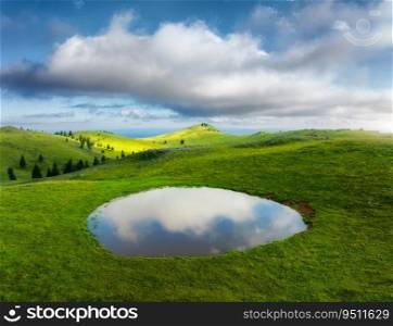 Aerial view of pond and green alpine meadows and hills at sunset in summer. Top drone view of mountain valley, green grass and sky with clouds reflected in water. Velika Planina, Slovenia. Landscape