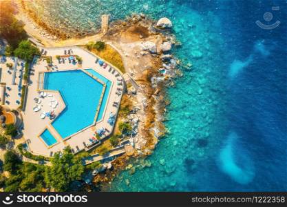 Aerial view of pool, sandy beach, stones, green trees, people and sea with transparent blue water at sunset. Top view of adriatic sea coast in summer. Landscape with azure water, rocks. Luxury resort