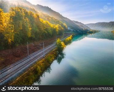 Aerial view of railroad near river in alpine mountains in fog at sunrise in autumn. Top view of rural railway station, lake, reflection in water, orange trees in fall. Railway station in Slovenia