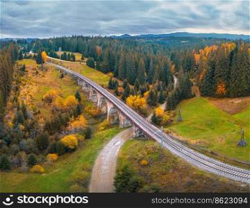 Aerial view of railway bridge, railroad, rural dirt road, green meadows, trees, hills and cloudy sky in fall. Top view of beautiful old viaduct at sunset in carpathian mountains in autumn in Ukraine