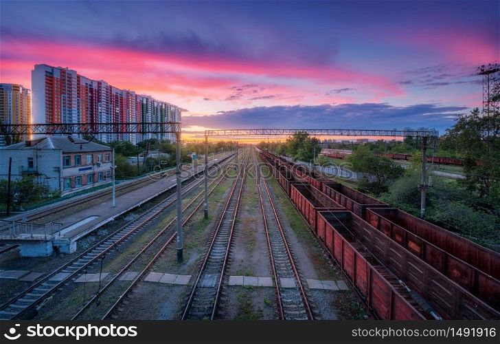 Aerial view of railway station with freight trains at colorful sunset in summer. Top view of railroad. Heavy industry. Landscape with train, buildings, sky with pink clouds at dusk. Transportation