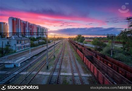 Aerial view of railway station with freight trains at colorful sunset in summer. Top view of railroad. Heavy industry. Landscape with train, buildings, sky with pink clouds at dusk. Transportation