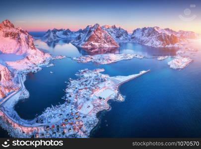 Aerial view of Reine at sunrise in winter. Top view of Lofoten islands, Norway. Landscape with blue sea, snowy mountains, high rocks, village with buildings, rorbu, blue sky, reflection in water