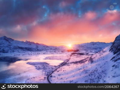 Aerial view of road, frozen sea coast, snow covered mountains, colorful sky with pink clouds at sunset in winter. Landscape frosty shore, snowy rocks, roadway. Lofoten islands, Norway. View from above