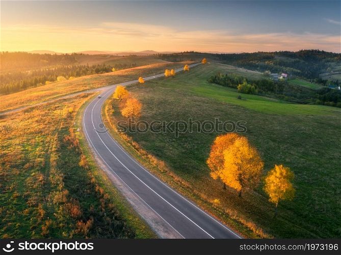 Aerial view of road, hills, green meadows and colorful trees at sunset in autumn. Top view of mountain rural road. Beautiful landscape with roadway, green grass, trees with orange foliage in fall