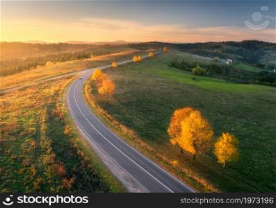 Aerial view of road, hills, green meadows and colorful trees at sunset in autumn. Top view of mountain rural road. Beautiful landscape with roadway, green grass, trees with orange foliage in fall