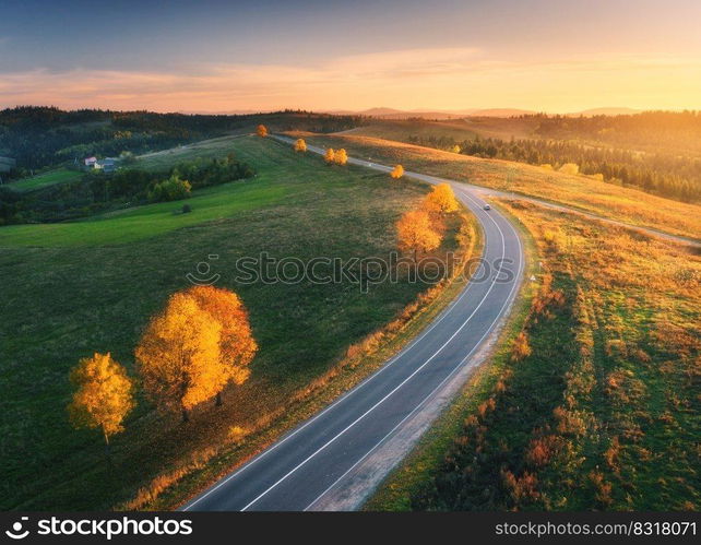 Aerial view of road, hills, green meadows and colorful trees at sunset in autumn. Top view of mountain rural road, golden sky. Beautiful landscape with roadway, grass, orange trees in fall. Highway