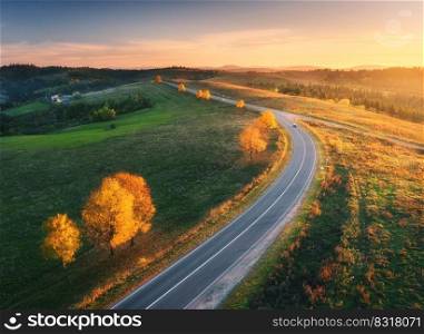 Aerial view of road, hills, green meadows and colorful trees at sunset in autumn. Top view of mountain rural road, golden sky. Beautiful landscape with roadway, grass, orange trees in fall. Highway