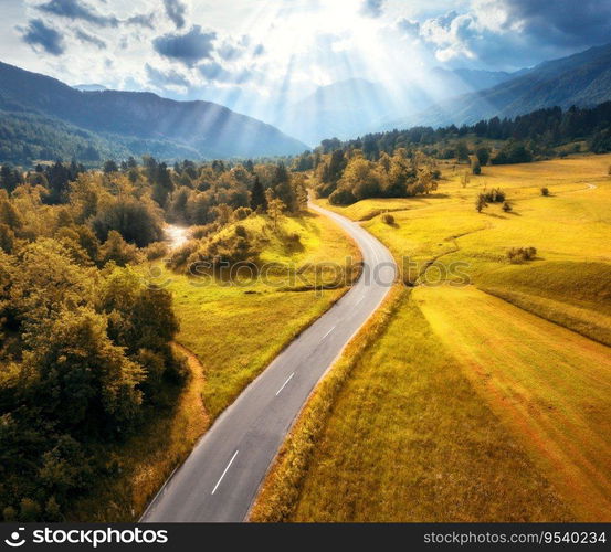 Aerial view of road in alpine meadows at sunset in autumn. Top view of rural road, mountains, forest in fall. Colorful landscape with country roadway, orange trees, hills, yellow grass in Slovenia