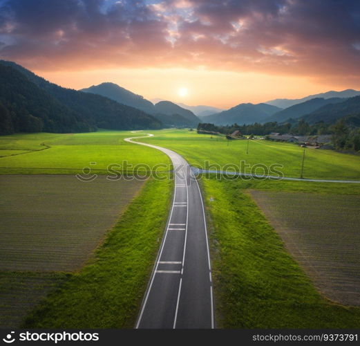 Aerial view of road in alpine mountain valley, green meadows at sunset in summer. Top drone view of country road. Colorful landscape with curved highway, hills, fields, grass, orange sky. Slovenia