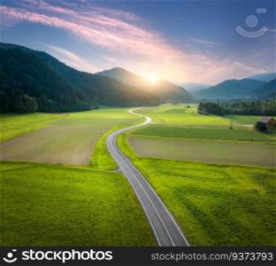 Aerial view of road in alpine mountain valley, green meadows at sunset in summer. Top drone view of country road. Colorful landscape with curved highway, hills, fields, grass, pink clouds. Slovenia
