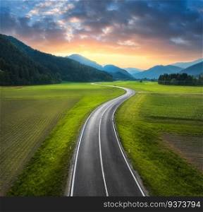Aerial view of road in alpine mountain valley, green meadows at sunset in summer. Top drone view of country road. Colorful landscape with curved highway, hills, fields, grass, orange sky. Slovenia