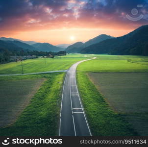 Aerial view of road in alpine mountain valley, green meadows at sunset in summer. Top drone view of country road. Colorful landscape with curved highway, hills, fields, grass, orange sky. Slovenia