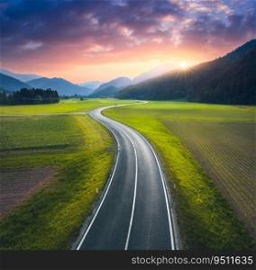 Aerial view of road in alpine mountain valley, green meadows at sunset in summer. Top drone view of country road. Colorful landscape with curved highway, hills, fields, grass, orange sky. Slovenia