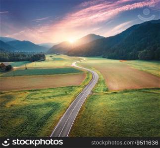 Aerial view of road in alpine mountain valley, green meadows at sunset in summer. Top drone view of country road. Colorful landscape with curved highway, hills, fields, grass, pink clouds. Slovenia