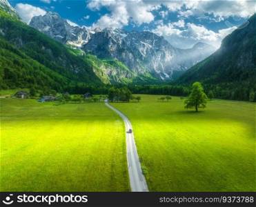 Aerial view of road in alpine mountains, green meadows, trees in summer. Top view of rural road. Colorful landscape with road, car, rocks, field, grass, sky, sunbeam, clouds. Logar valley, Slovenia