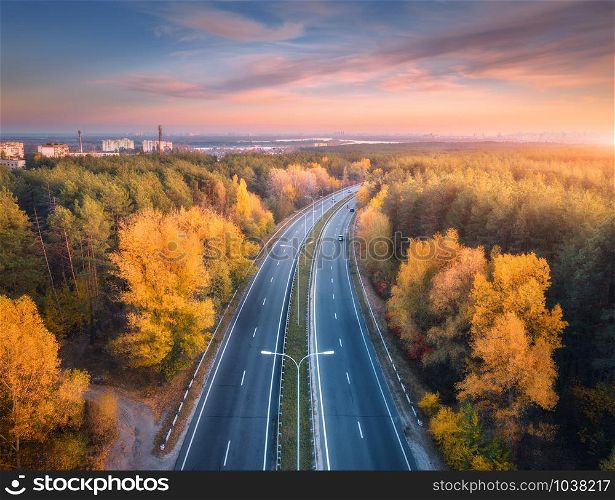 Aerial view of road in beautiful autumn forest at sunset. Colorful landscape with empty highway, trees with green and orange leaves, pink sky with clouds in fall. Top view of roadway. Autumn colors