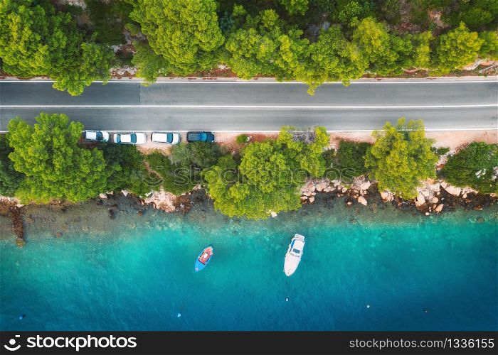 Aerial view of road in beautiful green forest and boats and yacht in the sea at sunset in summer. Colorful landscape with roadway, blue water, trees. Top view from drone of highway in Croatia. Travel