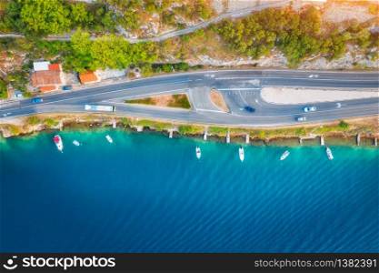 Aerial view of road in beautiful green forest and boats in the sea at sunset in summer. Colorful landscape with asphalt roadway, blue water, trees. Top view from drone of highway in Croatia. Travel. Aerial view of road in green forest and boats in the sea