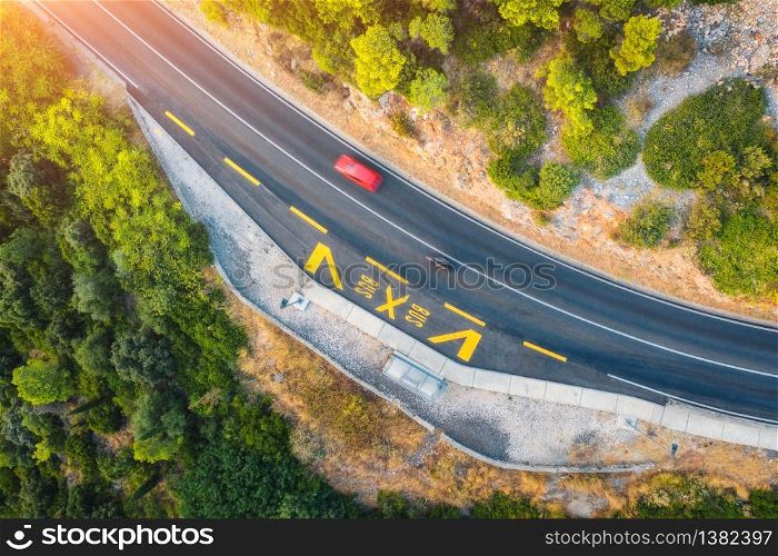 Aerial view of road in beautiful green forest at sunset in summer. Colorful landscape with red car on the roadway, trees in spring. Top view from drone of highway in Croatia. View from above. Travel. Aerial view of road in beautiful green forest at sunset in summer