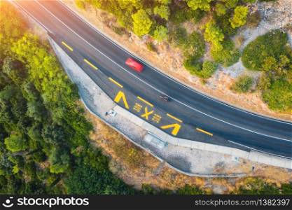 Aerial view of road in beautiful green forest at sunset in summer. Colorful landscape with red car on the roadway, trees in spring. Top view from drone of highway in Croatia. View from above. Travel. Aerial view of road in beautiful green forest at sunset in summer