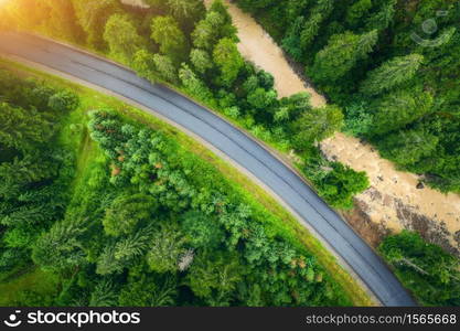 Aerial view of road in beautiful green forest at sunset in summer. Colorful landscape with roadway, pine trees, blurred car and river in Carpatian mountains. Top view of highway. Travel in Ukraine