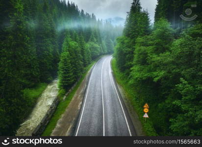 Aerial view of road in beautiful green forest in low clouds in rainy summer day. Colorful landscape with roadway in fog, pine trees, river in Carpatian mountains. Top view of road. Travel in Ukraine