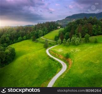 Aerial view of road in green alpine meadows at sunset in summer. Top view from drone of rural road, mountains, forest. Beautiful landscape with roadway, trees, hills, green grass, purple sky. Slovenia