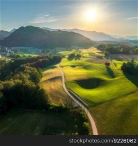 Aerial view of road in green alpine meadows at sunset in summer. Top view from drone of rural road, mountains, forest. Beautiful landscape with roadway, trees, hills, green grass, sky. Slovenia