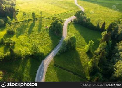 Aerial view of road in green meadows and hills at sunset in summer. Top view from drone of rural road, forest. Beautiful landscape with roadway, trees, green grass, sunlight in Slovenia. Nature