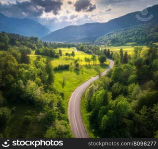 Aerial view of road in green meadows at summer sunny day. Top view from drone of rural road, mountains, forest. Beautiful landscape with roadway, sun rays, trees, hills, green grass, clouds. Slovenia