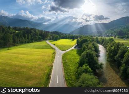 Aerial view of road in green meadows at sunset in summer. Top view from drone of rural road, river, mountains, forest. Beautiful landscape with roadway, sun rays, trees, hills, green grass. Slovenia