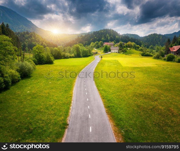 Aerial view of road in green meadows at sunset in summer. Top view from drone of rural road, mountains, forest. Beautiful landscape with roadway, sun rays, trees, hills, green grass, clouds. Slovenia