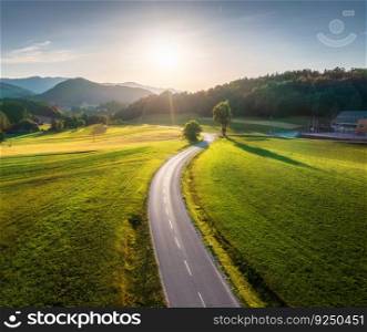 Aerial view of road in green meadows at sunset in summer. Top view from drone of rural road, mountains, forest. Beautiful landscape with roadway, trees, hills, fields, green grass, blue sky. Slovenia