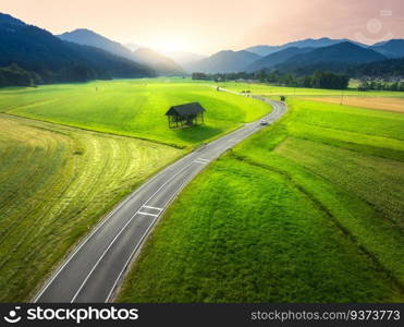 Aerial view of road in green meadows at sunset in summer. Top drone view of rural road, alpine mountains. Colorful landscape with curved highway, hills, fields, green grass, orange sky. Slovenia
