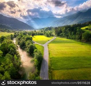 Aerial view of road in green meadows at sunset in summer. Top view from drone of rural road, river, mountains, forest. Beautiful landscape with roadway, sun rays, trees, hills, green grass in Slovenia