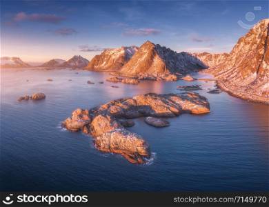 Aerial view of rocks in the sea, snowy mountains, blue sky with clouds at sunset. Travel in Lofoten Islands, Norway. Winter landscape with small islands in water, cliffs and waves. View from above