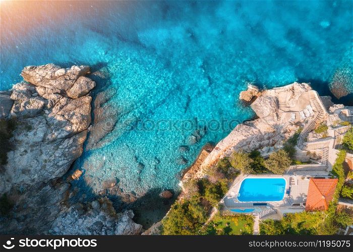Aerial view of rocky beach, pool, green trees and sea with transparent blue water at sunset. Coast of adriatic sea at sunset in summer. Top view. Landscape with azure water, rocks. Luxury resort