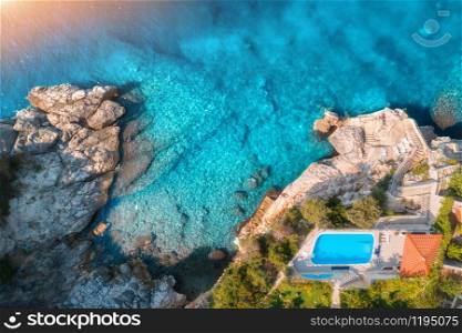 Aerial view of rocky beach, pool, green trees and sea with transparent blue water at sunset. Coast of adriatic sea at sunset in summer. Top view. Landscape with azure water, rocks. Luxury resort