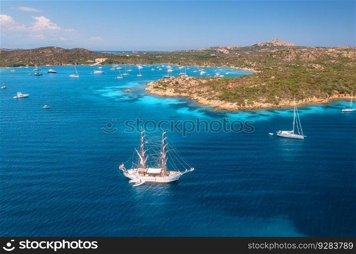 Aerial view of sail boat and luxury yachts on blue sea at sunset in summer. Sardinia, Italy. Tropical seascape with speed boats, yachts, sea bay, mountain, clear azure water, sky. Top view of ocean