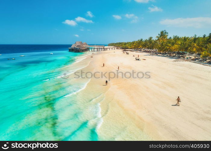 Aerial view of sandy beach of Indian Ocean at sunset. Summer holiday in Zanzibar, Africa. Tropical landscape with sea coast, palm trees, people, sand, clear blue water with waves. Top view. Seascape
