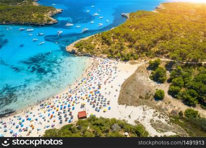 Aerial view of sandy beach with colorful umbrellas, swimming people, boats and yachts in sea bay, green trees and blue water at sunset in summer. Travel. Balearic islands, Spain. Top view. Landscape