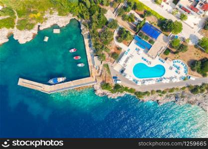 Aerial view of sea coast with clear blue water, boats, pier, hotel with swimming pool and green trees in summer at sunny day in Croatia. Top view of rocky beach, yachts, buildings and forest. Travel