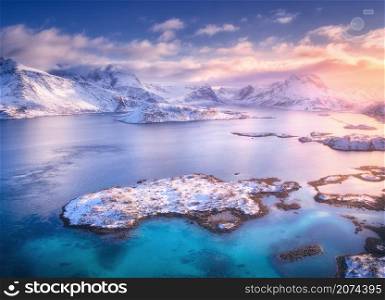 Aerial view of sea, rocks, islands and mountains in snow at sunset in winter. Lofoten islands, Norway. Landscape with snowy mountains, colorful sky with clouds. Scenery. Top view from drone. Nature