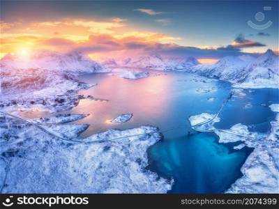 Aerial view of sea, snowy islands, mountains, road, blue sky at sunset in winter. Lofoten islands, Norway. Landscape with mountains and rocks in snow, reflection in water. Top view from drone. Nature